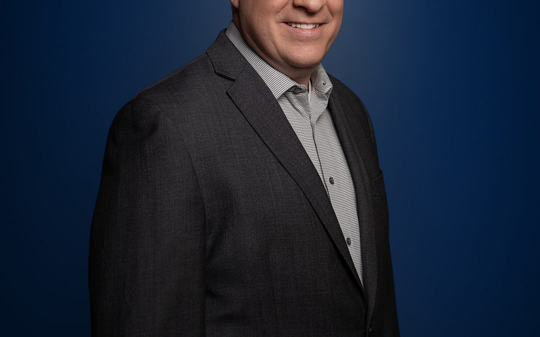 Scott Gray-Owen in a light grey shirt and charcoal blazer, smiling, against a dark blue background.