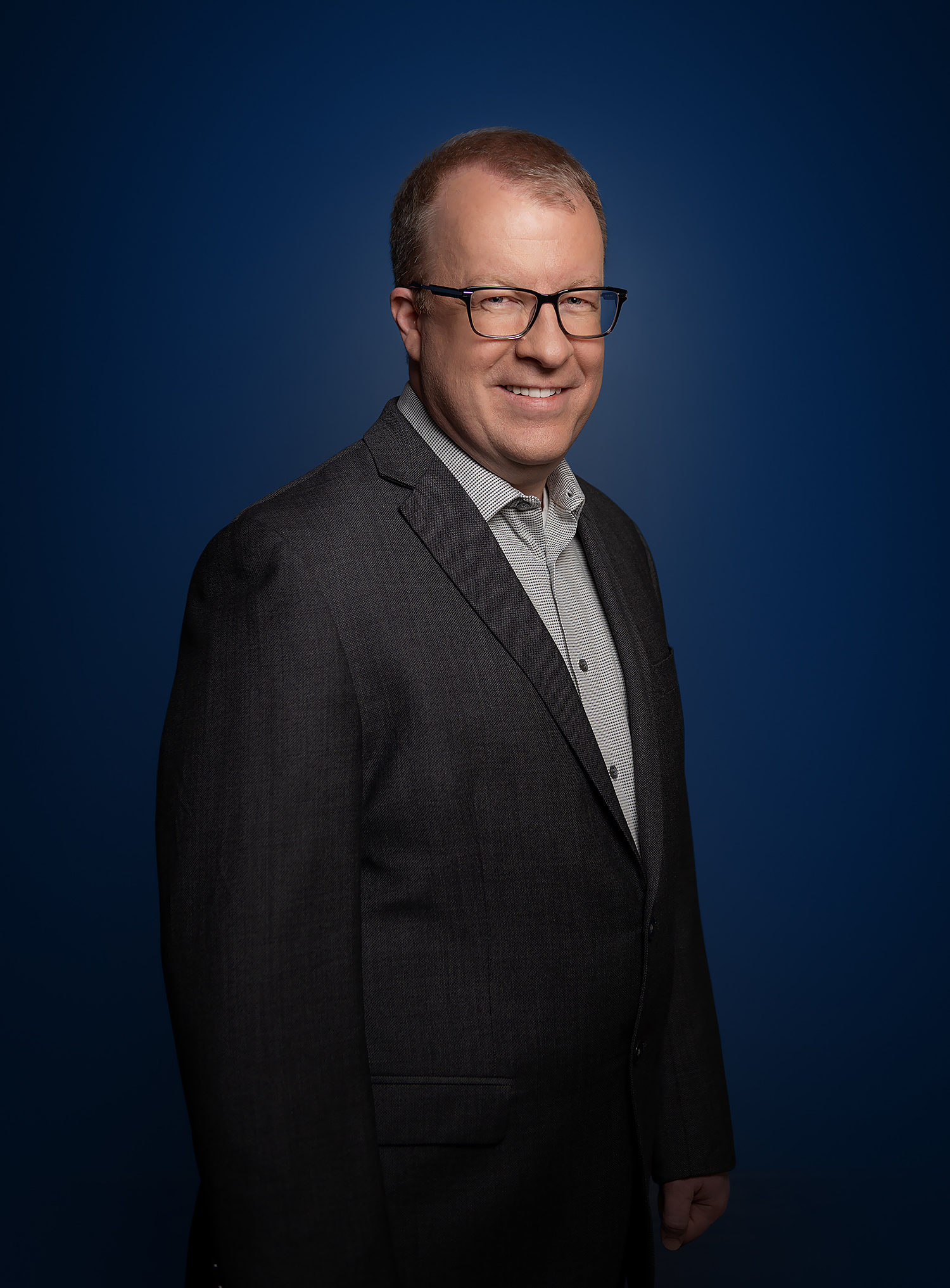 Scott Gray-Owen in a light grey shirt and charcoal blazer, smiling, against a dark blue background.