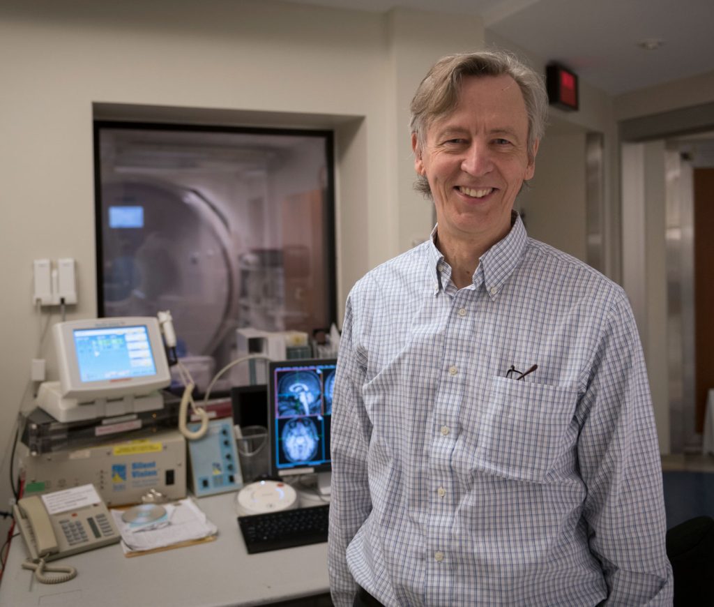 Dr. Kullervo Hynyen at Sunnybrook. He is wearing a light blue long-sleeved shirt, and in the background medical imaging equipment displays brain scans.